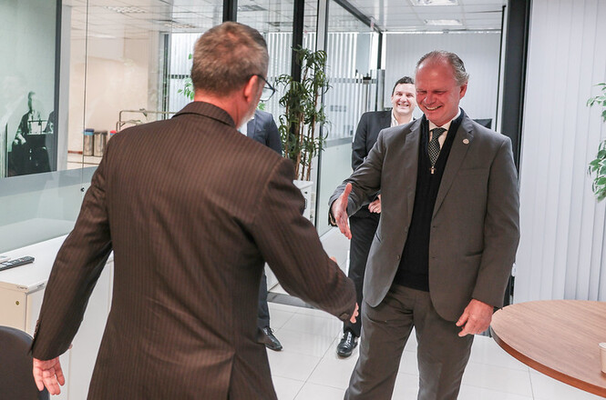 Anexo do usuário Foto horizontal: presidente do TJSC, desembargador Rubens Schulz, sorri ao cumprimentar com um aperto de mãos o oficial de justiça Paulo Roberto Costa, visto de costas, em ambiente institucional com divisórias de vidro. Ao fundo, dois homens acompanham o encontro.