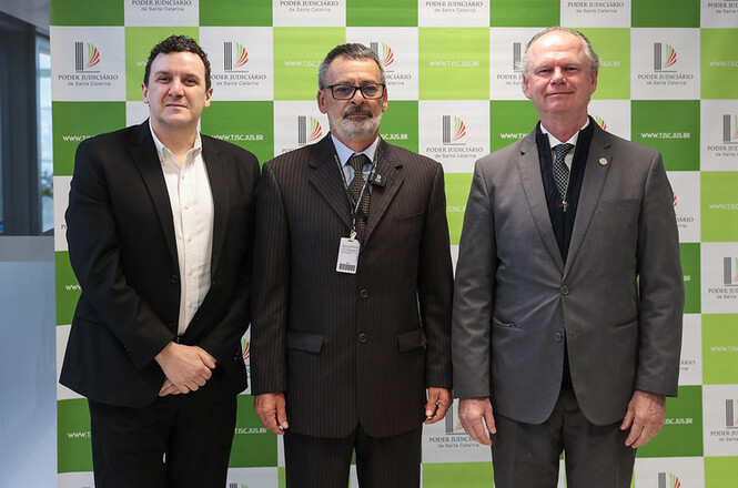 Foto horizontal: diretor-geral administrativo do TJSC, Alexsandro Postali, à esquerda, oficial de justiça Paulo Roberto Costa, ao centro, e presidente do TJSC, desembargador Rubens Schulz, à direita, posam para foto diante de painel institucional verde e branco do Poder Judiciário de Santa Catarina.