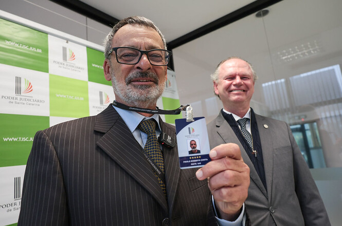 Foto horizontal: oficial de justiça Paulo Roberto Costa, de terno marrom e óculos, exibe seu crachá de identificação em primeiro plano, enquanto o presidente do TJSC, desembargador Rubens Schulz, sorri ao fundo, diante de painel institucional verde e branco.