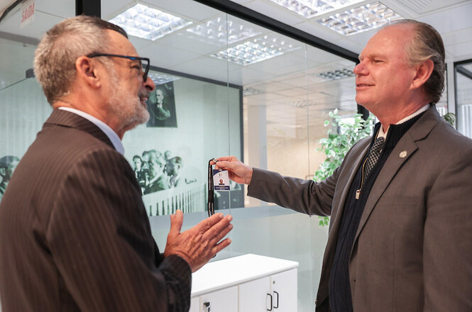 Foto horizontal: presidente do TJSC, desembargador Rubens Schulz, à direita, exibe o crachá de identificação do oficial de justiça Paulo Roberto Costa, que aparece de perfil, sorrindo e com as mãos próximas, durante encontro institucional em ambiente com divisórias de vidro.