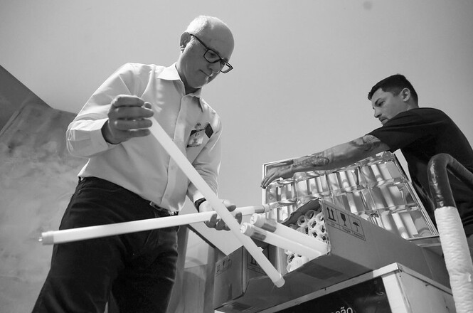 Horizontal: Participantes realizam atividade de reaproveitamento de materiais recicláveis, manipulando tubos e peças plásticas em ambiente interno de trabalho. Ao fundo, há caixas com materiais organizados, carrinho de transporte e equipamentos utilizados em ações voltadas à sustentabilidade e economia circular.