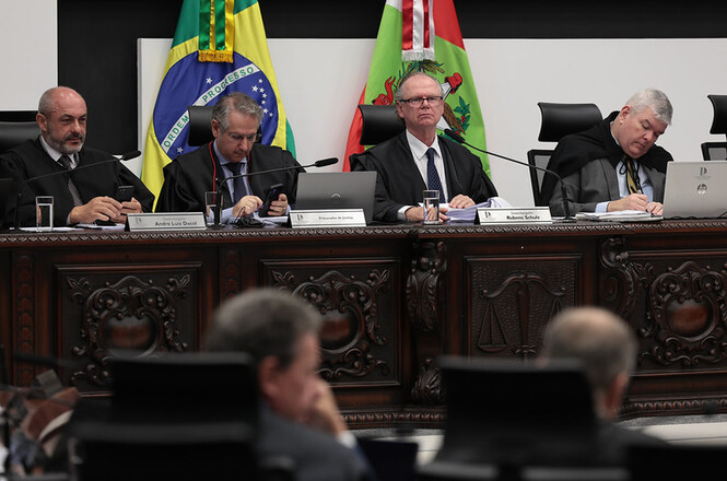 Foto colorida: Des. Rubens Schulz ao centro da mesa diretiva, plenário do Tribunal de Justiça, sessão judicial, acompanhando trabalhos com as bandeiras do Brasil e de Santa Catarina ao fundo em verde, vermelho e preto.
