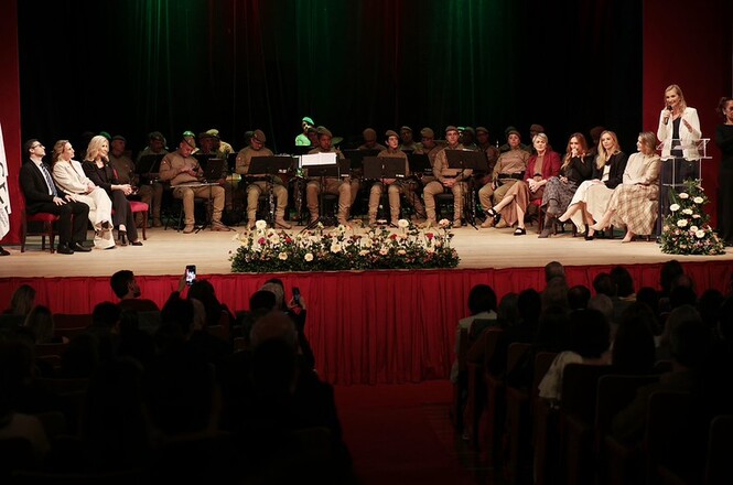 Foto horizontal: autoridades e banda militar em fardas cáqui e trajes formais pretos, bege e bordô sentam-se no palco com cortinas verde e vermelha enquanto mulher de blazer branco discursa ao púlpito durante cerimônia solene diante de plateia na penumbra.