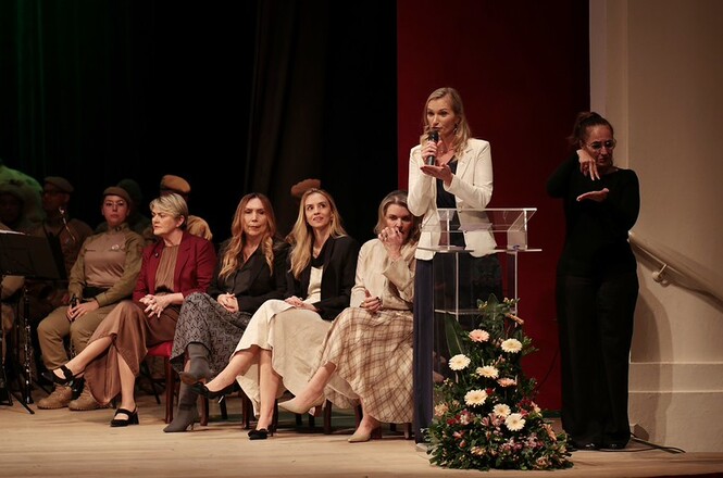 Foto horizontal: mulher em blazer branco discursa ao microfone em púlpito de acrílico ao lado de intérprete de Libras de preto e mulheres sentadas em poltronas com trajes bordô, preto, bege e farda cáqui em palco com cortinas escuras, coluna vermelha e arranjo de flores durante solenidade oficial.