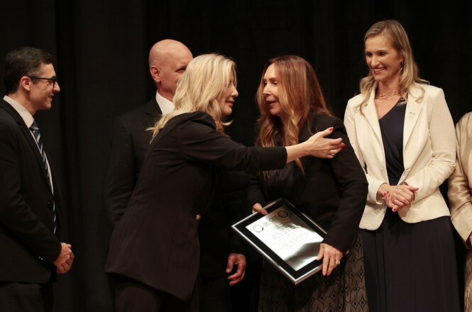 Foto horizontal: mulher em blazer preto cumprimenta com um abraço homenageada que segura placa solene, acompanhadas por autoridades em trajes preto, branco e azul-marinho em palco com fundo escuro durante cerimônia oficial de premiação.