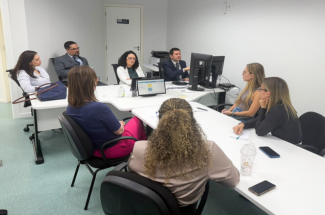 Foto horizontal: homem em terno azul-marinho aponta para a tela de monitor diante de grupo atento de participantes em trajes nas cores branco, cinza, preto, rosa e bege, sentados em cadeiras pretas ao redor de mesas modulares brancas em sala institucional.
