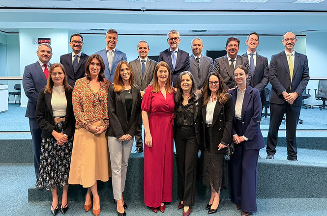 Foto horizontal: grupo de magistrados e autoridades em trajes formais nas cores azul-marinho, cinza, preto, vermelho, bege e branco posa alinhado e sorridente em dois planos sobre degraus com carpete verde-água, em auditório institucional iluminado e com paredes claras ao fundo.