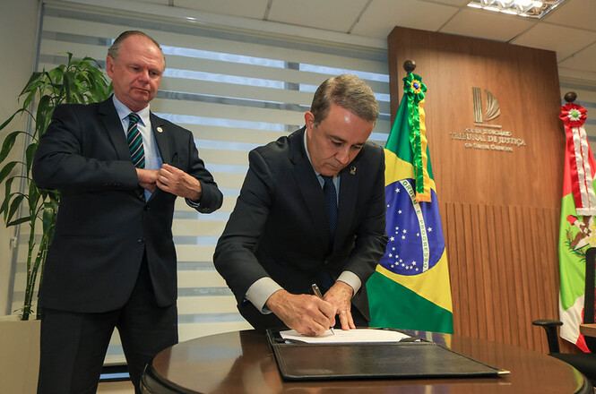 O presidente do TRE-SC, desembargador Carlos Roberto da Silva assina documento com caneta sobre pasta apoiada em mesa de madeira, ao lado da bandeira do Brasil, acompanhado pelo desembargador Rubens Schulz em pé, em gabinete institucional com o brasão do Tribunal de Justiça ao fundo durante ato solene.