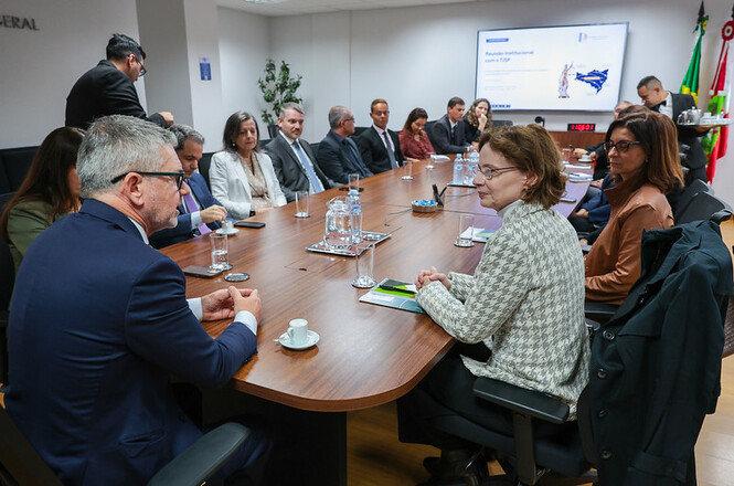 Foto horizontal: magistrados e servidores em trajes formais nas cores azul-marinho, bege, preto, cinza, marrom e branco dialogam sentados ao redor de longa mesa de madeira com copos d'água e xícaras de café, em sala de reuniões iluminada com tela ao fundo.