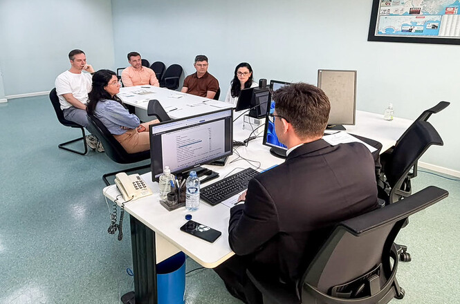 Foto horizontal: homem de terno preto em frente a monitor de computador dialoga com grupo de cinco participantes em trajes casuais nas cores branco, pêssego, marrom, azul-claro, bege e preto, sentados ao redor de mesas brancas em sala institucional.