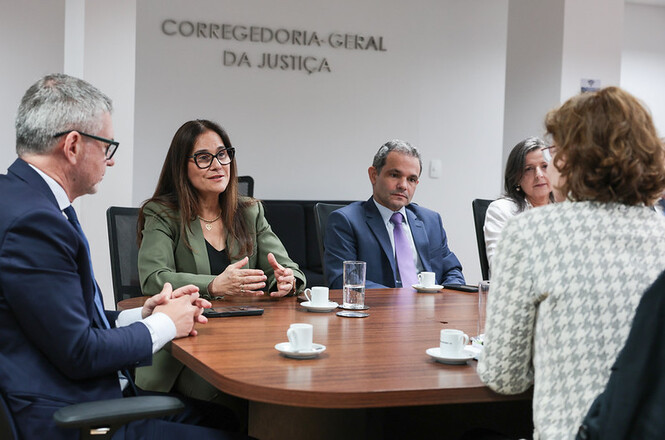 Foto horizontal: mulher de blazer verde-oliva e óculos gesticula e dialoga com magistrados e servidores em trajes formais nas cores azul-marinho, lilás, branco, cinza e preto, sentados ao redor de mesa de madeira oval com xícaras de café em sala institucional com a inscrição "Corregedoria-Geral da Justiça" na parede clara ao fundo.