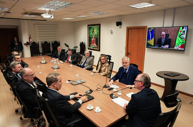 Foto horizontal: grupo de magistrados e servidores em trajes formais nas cores azul-marinho, cinza, preto, bege e vermelho senta-se ao redor de longa mesa retangular de madeira clara com microfones de mesa, copos e xícaras de café, em sala de reuniões institucional.