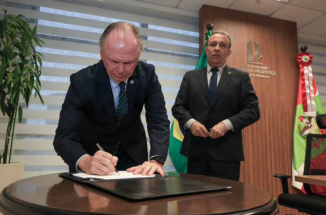 O presidente do TJSC, desembargador Rubens Schulz assina documento com caneta sobre pasta apoiada em mesa redonda de madeira, acompanhado pelo desembargador Carlos Roberto da Silva em pé, em gabinete institucional com o brasão do Tribunal de Justiça e bandeiras oficiais com detalhes em verde, amarelo e vermelho ao fundo durante ato solene.