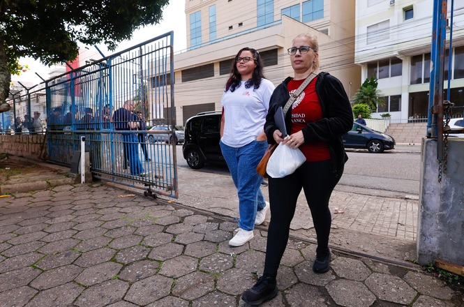 Foto horizontal: mulheres chegam ao local de prova em área externa, movimentação ao fundo, clima de expectativa e participação no exame.