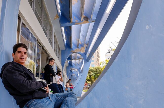 Foto horizontal: pessoas aguardam em área externa de local de prova, apoiadas em estrutura, clima de expectativa e intervalo do exame.