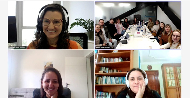 Foto horizontal: tela de videoconferência mostra três mulheres conectadas remotamente e um grupo reunido presencialmente em sala de reunião.