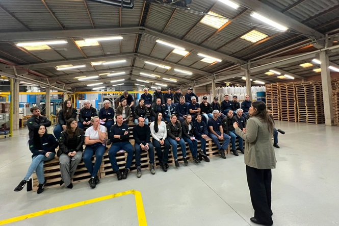 Foto horizontal: trabalhadores acompanham palestra em ambiente industrial, com palestrante à frente do grupo.