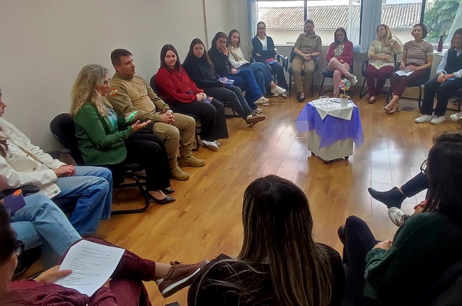 Imagem horizontal mostra mulheres e policial militar reunidos em roda de conversa durante atividade do Agosto Lilás.