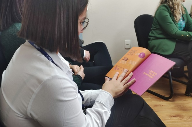 Imagem horizontal mostra mulher sentada lendo cartilha informativa durante atividade do Agosto Lilás.