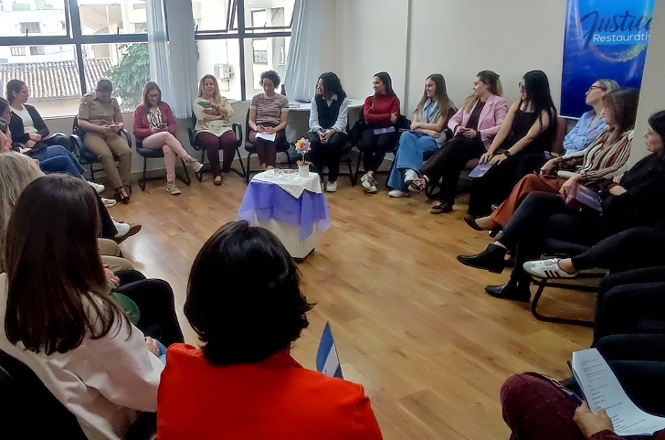 Imagem horizontal mostra mulheres e policial militar reunidos em roda de conversa durante atividade do Agosto Lilás.
