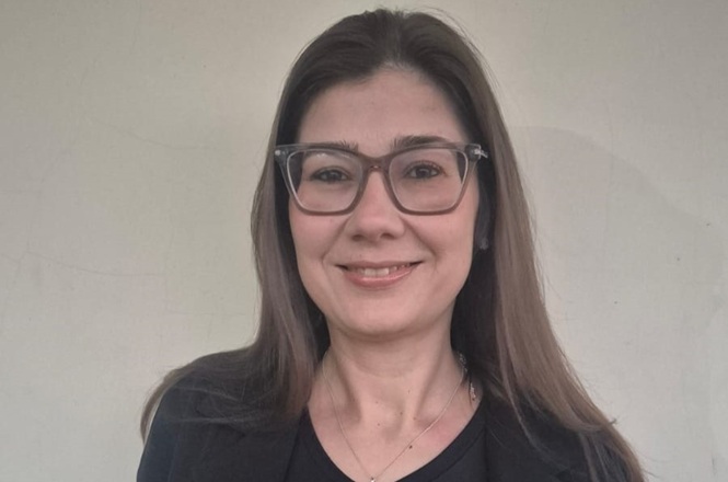 Foto horizontal: psicóloga Amanda Bicca posa sorridente para retrato, usando óculos e olhando em direção à câmera.