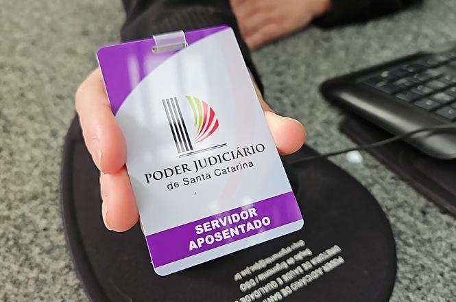 Foto horizontal: mão segura e exibe crachá funcional em primeiro plano com detalhes em roxo, branco, preto, verde e vermelho, apresentando o logotipo e a inscrição "Poder Judiciário de Santa Catarina - Servidor Aposentado", sobre mesa de trabalho institucional.