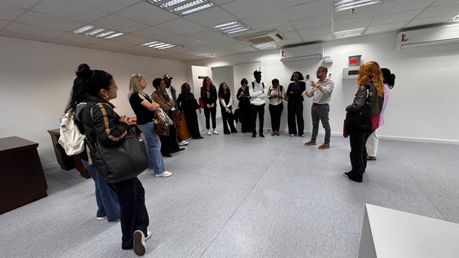 Foto horizontal: homem gesticula ao falar para grupo de estudantes reunido em círculo em ampla sala branca