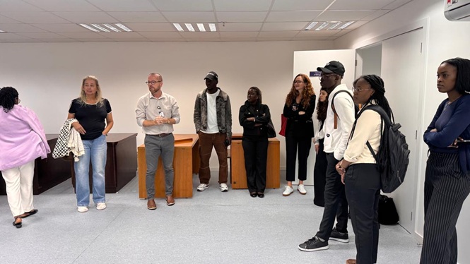 Foto horizontal: homem de camisa clara e crachá conversa com grupo de estudantes em pé numa sala ampla com mesas de madeira ao fundo, durante visita técnica ao Arquivo Central do TJSC.