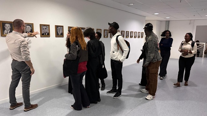 Foto horizontal: homem aponta para galeria de retratos históricos na parede enquanto orienta estudantes em fila