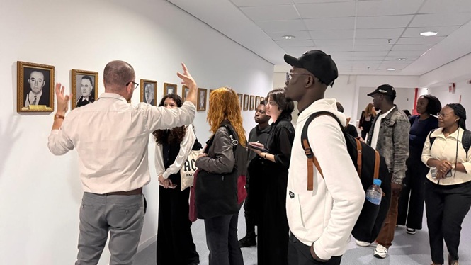 Foto horizontal: homem gesticula enquanto explica galeria de retratos emoldurados na parede para grupo de estudantes