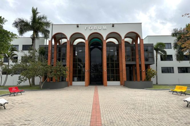 Foto horizontal: fachada do Fórum de Tubarão, com arcos de tijolos, palmeiras e bancos coloridos na área externa.