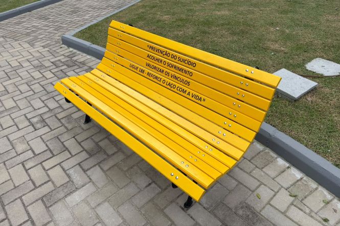 Foto horizontal: banco amarelo exibe mensagens de prevenção ao suicídio e valorização da vida em área externa.