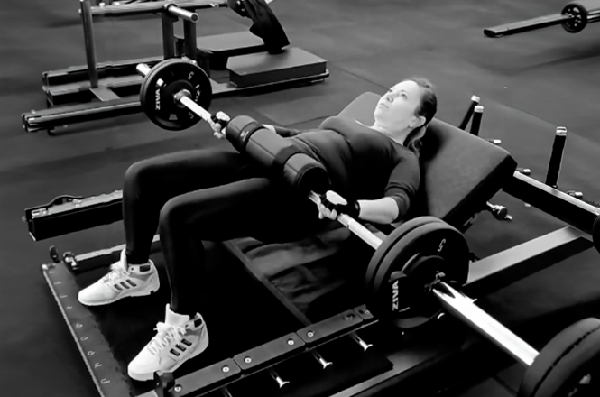 Foto horizontal: mulher realiza exercício de força com barra e pesos em academia, em imagem em preto e branco.