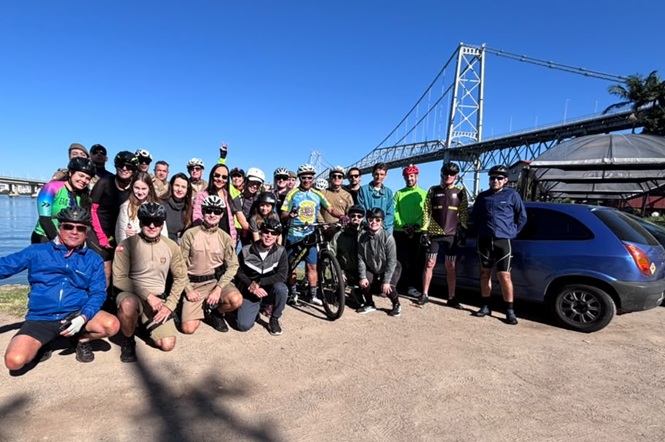 Foto horizontal: grupo de ciclistas posa para registro coletivo às margens da Baía Sul, com a Ponte Hercílio Luz ao fundo.