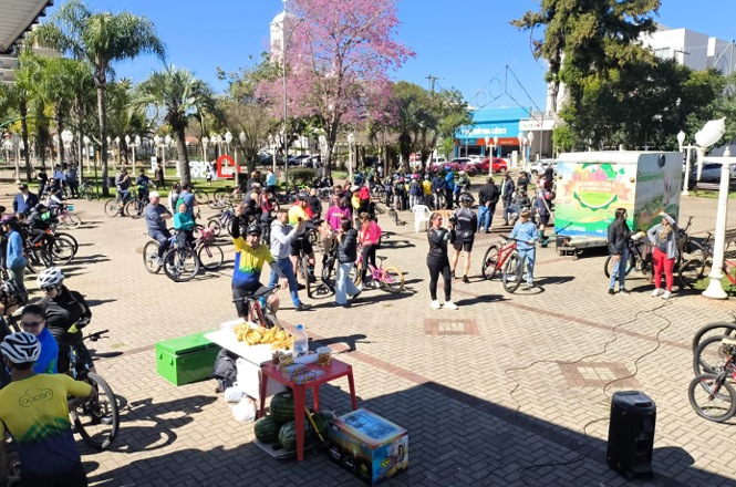 Foto horizontal: grande grupo de ciclistas se reúne com bicicletas em praça durante passeio coletivo, com ponto de apoio montado no local.