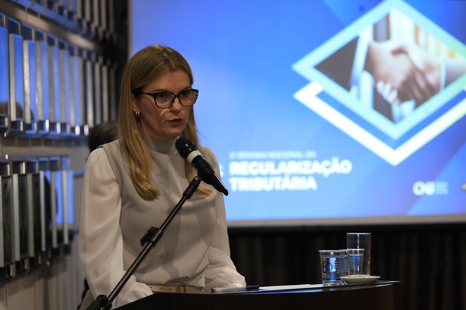 Foto horizontal: juíza Fabíola Duncka Geiser discursa ao microfone durante evento sobre regularização tributária, com painel temático ao fundo.