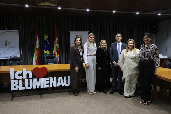Foto horizontal: magistrados e magistradas posam para foto no plenário, diante de bandeiras e do letreiro “Ich ❤️ Blumenau”.