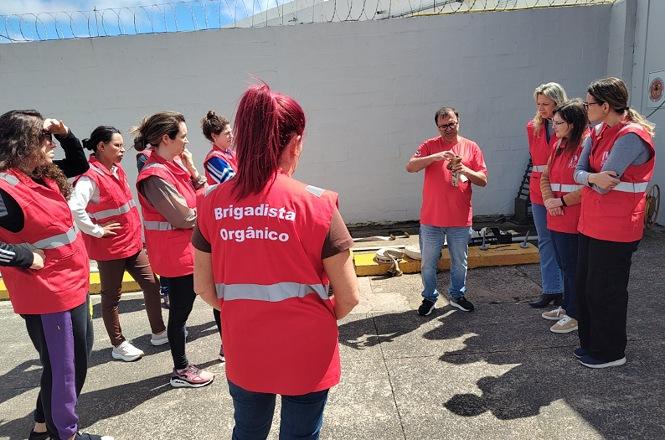 Foto horizontal: instrutor de camiseta vermelha orienta grupo de brigadistas durante treinamento prático em área externa, com participantes usando coletes vermelhos identificados como “Brigadista Orgânico” e acompanhando a demonstração.