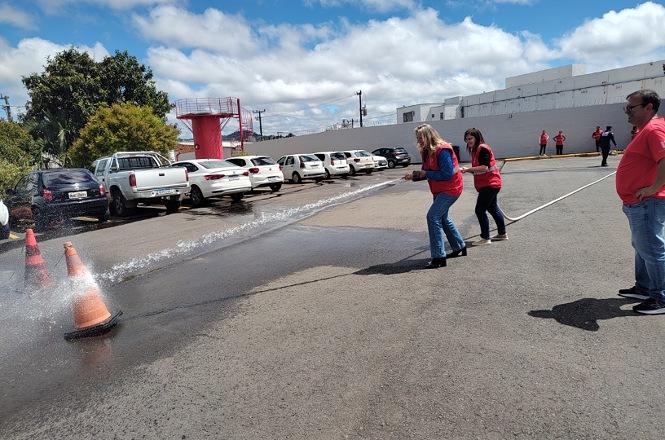 Foto horizontal: duas brigadistas com coletes vermelhos participam de treinamento prático de combate a incêndio, manuseando mangueira que lança jato de água em área externa, próxima a cones de sinalização, enquanto instrutor acompanha a atividade.