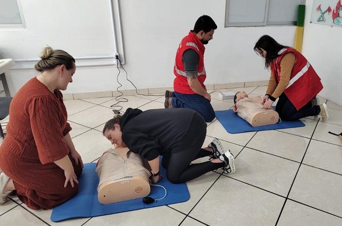 Foto horizontal: participantes de treinamento de primeiros socorros praticam manobras de reanimação cardiopulmonar (RCP) em manequins posicionados sobre colchonetes azuis, enquanto dois brigadistas com coletes vermelhos acompanham a atividade.