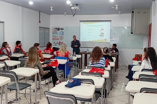 Foto horizontal: instrutor do Corpo de Bombeiros, de uniforme azul-escuro, conduz treinamento para participantes sentados em carteiras escolares, alguns com coletes vermelhos, enquanto uma apresentação é projetada na parede da sala de aula.