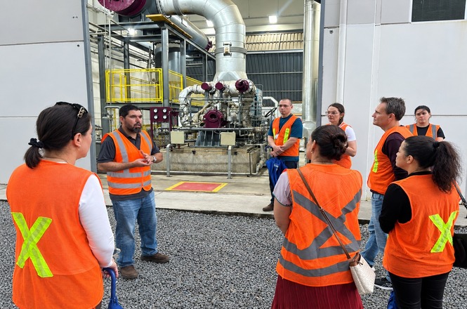Foto horizontal: grupo com coletes laranja participa de visita técnica em área industrial, diante de máquinas e tubulações.