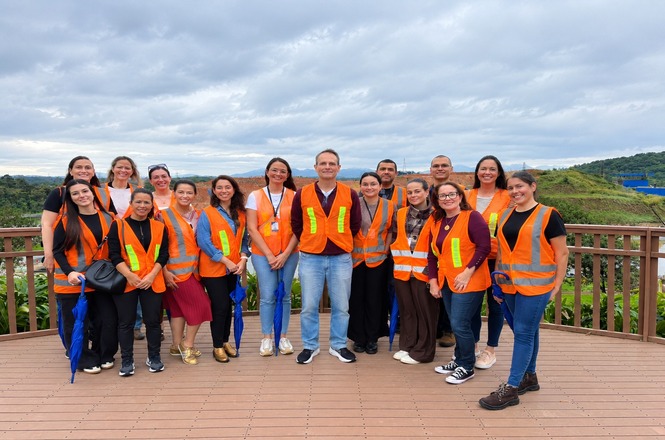 Foto horizontal: grupo com coletes laranja posa em área externa durante visita técnica, com vegetação ao fundo.
