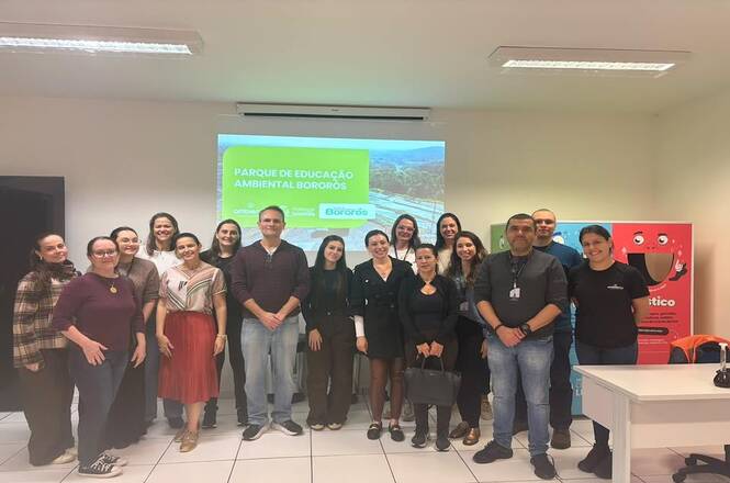 Foto horizontal: grupo posa após atividade no Parque de Educação Ambiental Bororós, com apresentação projetada ao fundo.