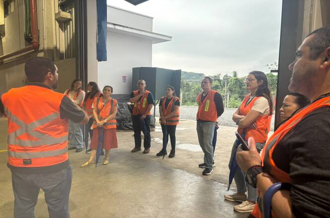 Foto horizontal: participantes de coletes laranja ouvem orientações durante visita técnica em instalação industrial.