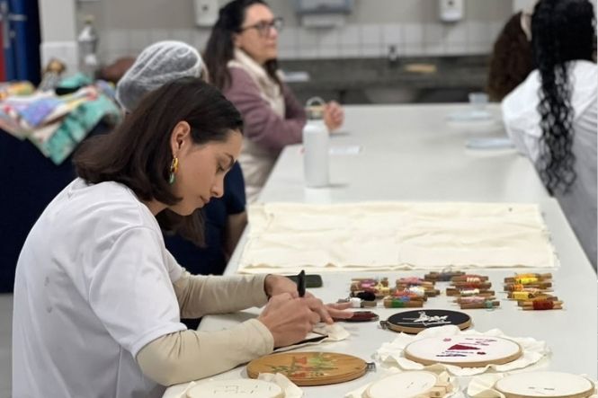 Foto horizontal: mulher de branco borda tecido à mesa, cercada por linhas coloridas, bastidores e outras participantes.