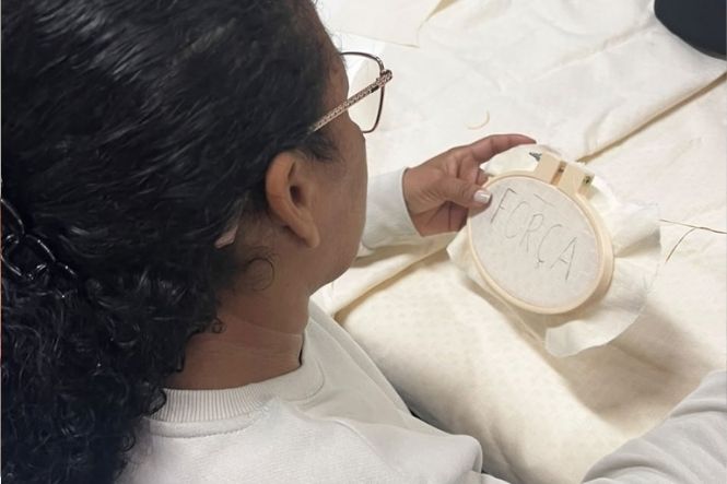 Foto horizontal: participante de branco segura bastidor com tecido e letras traçadas durante oficina de bordado.