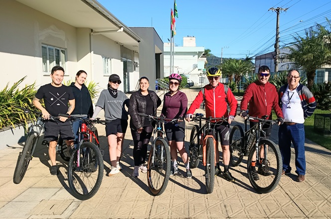 Foto horizontal: sete pessoas posam para registro coletivo, cinco delas ao lado de bicicletas, em frente a prédio com bandeiras hasteadas.
