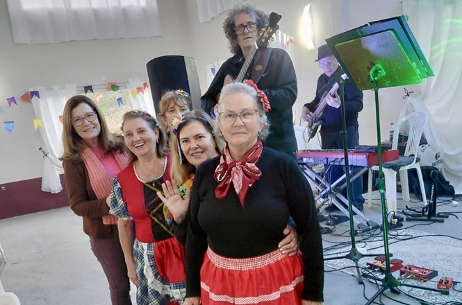 Foto horizontal: Servidoras posam com trajes típicos durante festa julina, enquanto músicos se apresentam ao fundo.