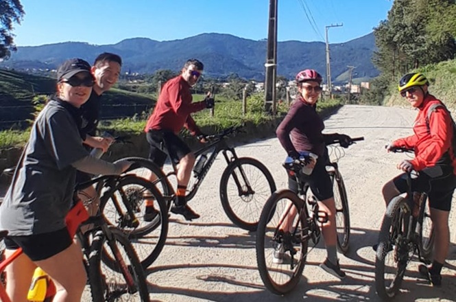 Foto horizontal: cinco ciclistas posam com suas bicicletas em estrada, com montanhas e vegetação ao fundo.